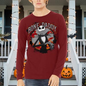 Może przedstawiać: Czerwona koszula z długim rękawem z grafiką Jacka Skellingtona z filmu Koszmar przed świętami. Grafika zawiera napis "Bone Daddy".