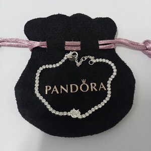 Op de afbeelding: Een zilveren armband met talrijke kleine, heldere edelstenen, liggend op een zwarte fluwelen zak. De zak heeft het woord "PANDORA" in het wit gedrukt. De armband is verbonden met een kleine sluiting.