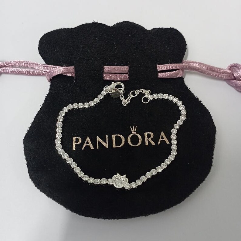 Op de afbeelding: Een zilveren armband met talrijke kleine, heldere edelstenen, liggend op een zwarte fluwelen zak. De zak heeft het woord "PANDORA" in het wit gedrukt. De armband is verbonden met een kleine sluiting.