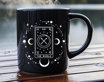 Puede incluir: Taza de cerámica negra con un diseño de carta de tarot blanco, con las palabras "THE WORLD". El diseño incluye lunas crecientes, estrellas y símbolos astrológicos. Sale vapor de la taza.