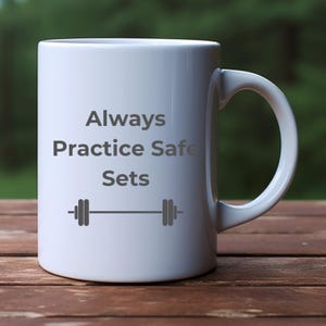 Könnte beinhalten: Weiße Keramiktasse mit einem grauen Design, auf dem "Always Practice Safe Sets" und eine Hantelgrafik abgebildet sind.