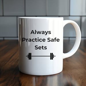 Könnte beinhalten: Weiße Keramik-Tasse mit dem Text "Always Practice Safe Sets" in Schwarz und einer Hantel-Grafik. Die Tasse hat einen gebogenen Henkel und steht auf einer Holzoberfläche.