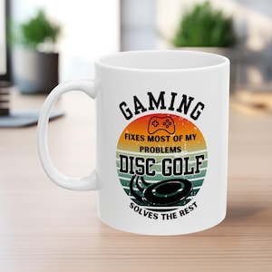 May include: White ceramic mug with a black handle featuring the text "GAMING fixes most of my problems, DISC GOLF solves the rest." The design includes a controller graphic and a disc golf disc.