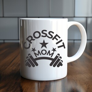 May include: White ceramic mug featuring the text "CROSSFIT MOM" in black. The design includes a star and a barbell graphic. The mug has a curved handle. It is set on a wooden surface.