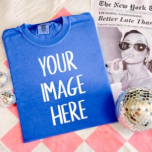 May include: A blue t-shirt with the text "YOUR IMAGE HERE" printed in white on the front. The t-shirt is folded and lying on a pink and white checkered surface.