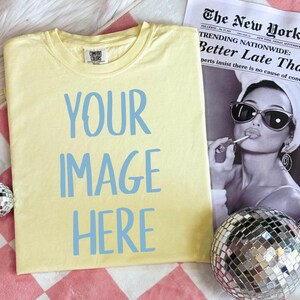 Puede incluir: Una camiseta amarilla con el texto "YOUR IMAGE HERE" impreso en azul claro. La camiseta está sobre una superficie a cuadros rosa y blanca. Hay una bola de discoteca plateada y un periódico con el titular "Better Late Than Never" en el fondo.