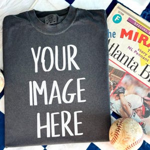 May include: A dark gray t-shirt with the text "YOUR IMAGE HERE" printed in white on the front. The t-shirt is lying on a white and blue checkered surface.