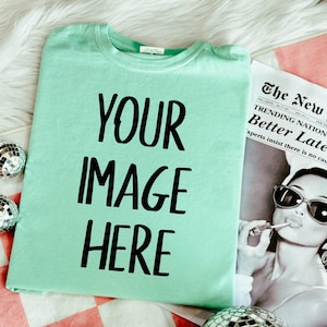 Może przedstawiać: Jasnozielona koszulka z czarnym napisem "YOUR IMAGE HERE". Koszulka leży na białej, puszystej powierzchni obok gazety z nagłówkiem "The New". Obecne są również dekoracyjne kule dyskotekowe.