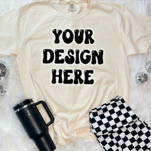 May include: A cream colored t-shirt with the text "YOUR DESIGN HERE" in black bubble letters.