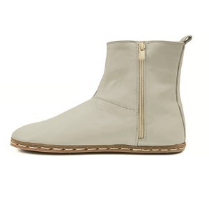 May include: A pale grey ankle boot featuring a side zip and stitched sole. The boot has a rounded toe and a pull tab. The zip is gold in colour.