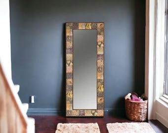 May include: A tall, rectangular mirror with a decorative mosaic frame. The frame features a repeating pattern of squares in shades of gold, brown, and beige. The mirror is positioned against a dark gray wall, with a basket and rug visible.