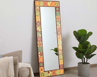 May include: A full-length mirror with a decorative frame. The frame is made of dark wood and features a mosaic pattern of colorful square tiles in shades of orange, green, yellow, and red. The mirror is leaning against a white wall.