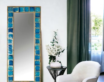 May include: A large rectangular mirror with a gold frame and a border of square, turquoise tiles. The mirror is placed against a white wall, with a small table, flowers, and a white armchair nearby.