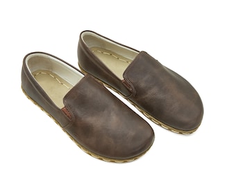 May include: A pair of brown leather slip-on shoes. The shoes have a rounded toe, a stitched sole, and a small elastic panel on the side. The interior is a light tan color. The shoes are displayed on a white background.