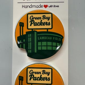 Puede incluir: Dos posavasos redondos para coche con el logotipo de los Green Bay Packers y el estadio Lambeau Field. Los posavasos tienen un fondo naranja con un dise&ntilde;o de estadio verde y negro. El texto "Car Coasters" y "Handmade with love" est&aacute; en la parte superior.