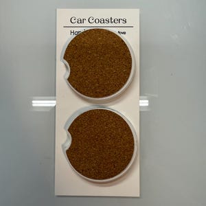 Puede incluir: Dos posavasos para coche con centro de corcho y borde de pl&aacute;stico transparente, expuestos en una tarjeta blanca. La tarjeta dice "Car Coasters" en la parte superior. Los posavasos est&aacute;n dise&ntilde;ados para caber en el portavasos de un coche.