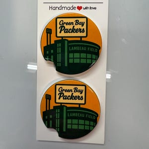 Puede incluir: Dos posavasos para coche con el logotipo de los Green Bay Packers y el estadio Lambeau Field sobre un fondo naranja. Los posavasos son redondos y tienen una peque&ntilde;a muesca. El texto "Car Coasters" y "Handmade with love" est&aacute; impreso.