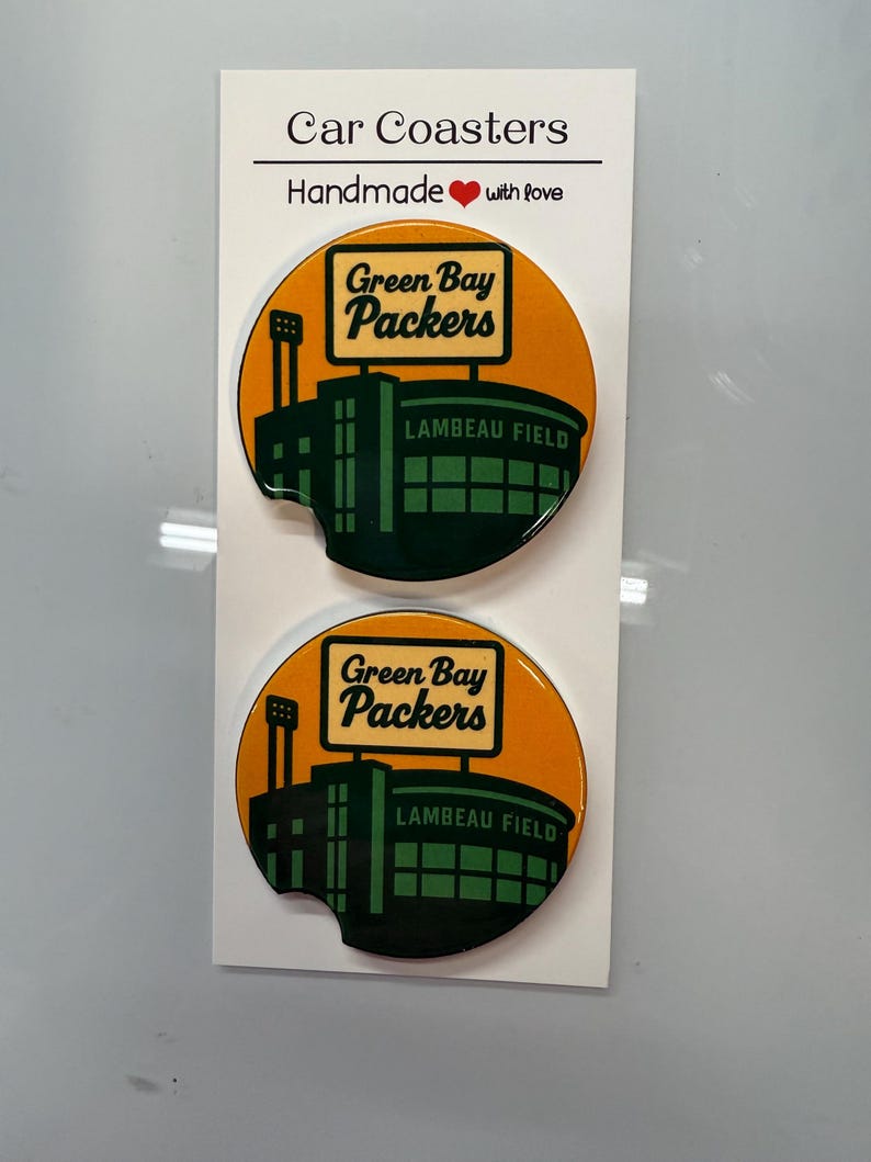 Puede incluir: Dos posavasos para coche con el logotipo de los Green Bay Packers y el estadio Lambeau Field sobre un fondo naranja. Los posavasos son redondos y tienen una peque&ntilde;a muesca. El texto "Car Coasters" y "Handmade with love" est&aacute; impreso.