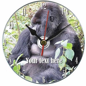 Puede incluir: Reloj de pared redondo con una imagen de gorila. La esfera del reloj tiene manecillas negras, un segundero rojo y números blancos. El texto "Your text here" está impreso en la esfera. El fondo muestra follaje verde.