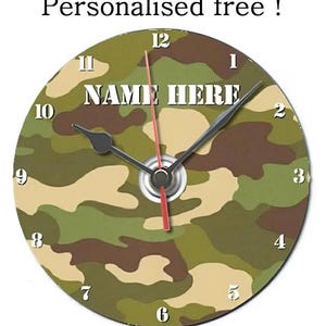 Puede incluir: Reloj de pared redondo con un diseño de camuflaje en tonos verdes, marrones y beige. La esfera del reloj muestra el texto "NAME HERE" en blanco. Agujas negras para horas y minutos, y un segundero rojo.