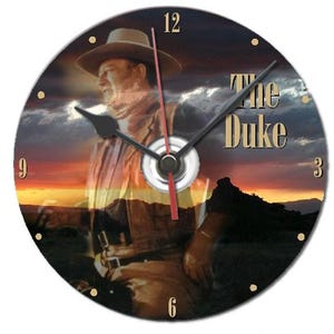 Peut inclure: Horloge murale ronde avec l'image d'un cowboy portant un chapeau sur fond de coucher de soleil. Le cadran de l'horloge affiche le texte "The Duke" en or, des aiguilles noires et des repères horaires dorés.