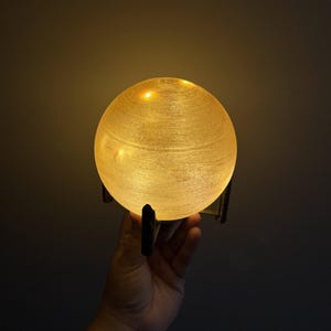 Peut inclure: Une lampe sphérique lumineuse avec une surface texturée, maintenue par un support sombre à trois pieds. La lampe émet une lumière dorée et chaude, créant une lueur ambiante douce. Un élément de décoration pour la maison.
