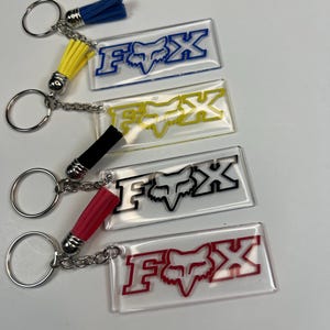 Könnte beinhalten: Vier Schlüsselanhänger mit dem "FOX"-Logo in Blau, Gelb, Schwarz und Rot. Jeder Schlüsselanhänger hat ein rechteckiges, transparentes Acrylschild mit dem Logo, einen Metallring und eine Quaste in passender Farbe. Zubehör.