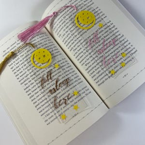 Puede incluir: Marcadores de acrílico transparente con diseños de luna y estrellas amarillas, y borlas rosas y doradas. Un marcador tiene las palabras "sleepless" y "bless" en escritura dorada.