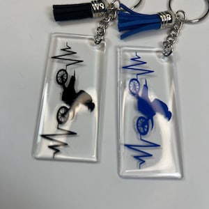May include: Two rectangular keychains with a motorcycle design and a heartbeat graphic. One keychain has a black motorcycle design and a black tassel, the other has a blue motorcycle design and a blue tassel. Both have silver key rings.