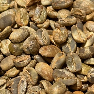 Puede incluir: Primer plano de un montón de granos de café crudos y sin tostar. Los granos presentan varios tonos de verde y marrón, con grietas y texturas visibles. La imagen destaca el estado natural y sin procesar de los granos de café.