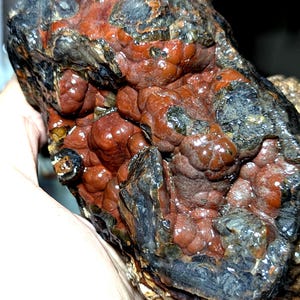 May include: A raw mineral specimen featuring a combination of dark, metallic charcoal-coloured crystalline structures and smooth, rounded, reddish-brown botryoidal formations. The surface exhibits a high-gloss, reflective texture throughout.