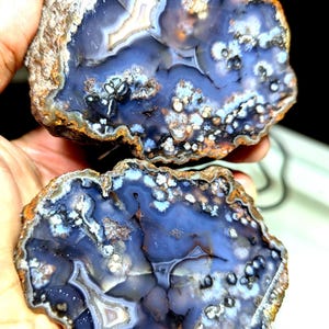 Può includere: Due metà di una geode di agata lucidata, che mostra un interno blu intenso con motivi bianchi, grigi e marroni. I bordi esterni sono di un marrone terroso ruvido. La geode è tenuta tra le mani di una persona, evidenziando la bellezza naturale della pietra.