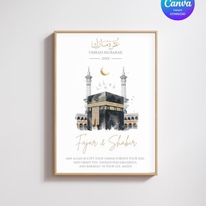 May include: A framed art print featuring a watercolor illustration of the Kaaba in Mecca. The artwork includes Arabic calligraphy and English text that reads "Umrah Mubarak" and "Fajar & Shabir". The print has a gold frame and a white background.