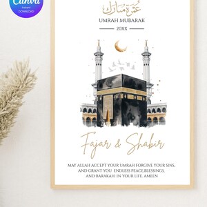 May include: A framed print with a watercolor illustration of the Kaaba in Mecca, with gold accents and Arabic calligraphy. The text reads "Umrah Mubarak" and "Fajar & Shabir," with a blessing below. The print is in a light wood frame.