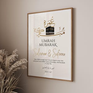Op de afbeelding: Een ingelijste print met de woorden "UMRAH MUBARAK" in het Engels en Arabische kalligrafie. De print toont een afbeelding van de Kaaba en bevat de namen "Suleman & Safeena", samen met een gebed.