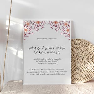 Puede incluir: Una impresión enmarcada con el texto "DUA FOR PROTECTION" en inglés y escritura árabe. El marco blanco está decorado con detalles florales en tonos rosa y naranja. Hay un posavasos tejido al lado del marco.