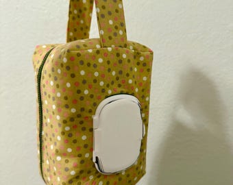 May include: A fabric wipe dispenser with a handle, featuring a mustard-colored background with pink, white, and brown polka dots. The dispenser has a white plastic opening for easy access to wipes. The zipper is green.