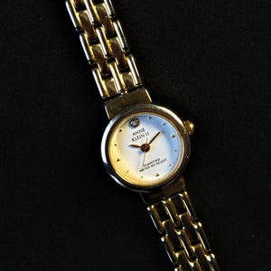 Puede incluir: Un reloj de pulsera de tono dorado con una esfera blanca y agujas negras. La esfera del reloj tiene un pequeño diamante en la posición de las 12 en punto y el texto "Anne Klein II Diamond Water 100 Resist".