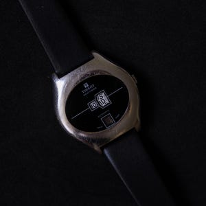 May include: A Tissot Newtimer automatic wristwatch with a silver-tone metal case and a black leather strap. The black dial features white text reading Tissot Newtimer and Automatic, with a unique digital-style display showing the numbers 15 and 10.