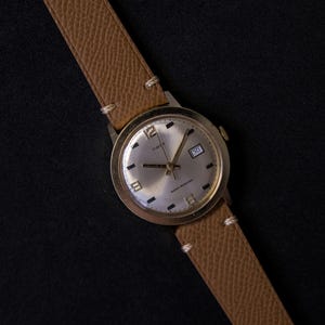 Könnte beinhalten: Eine Timex Armbanduhr mit rundem silberfarbenem Zifferblatt, goldfarbenen Stundenmarkierungen und einem Datumsfenster, das 30 anzeigt. Die Uhr hat ein strukturiertes, beiges Lederarmband mit weißen Nähten und ein goldfarbenes Metallgehäuse.