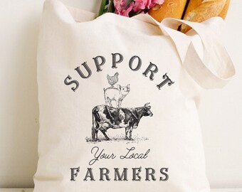 May include: A cream-colored canvas tote bag featuring a black graphic print of a cow, a pig, and a chicken stacked vertically. The text reads SUPPORT Your Local FARMERS. The bag is filled with two golden-brown baguettes and pink snapdragon flowers.