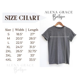May include: A gray t-shirt hangs on a wooden hanger, with measurements indicated by arrows. A size chart displays shirt dimensions in inches: S (18.5" width, 27" length), M (20.5" width, 28.5" length), L (22.5" width, 30" length), and more. The text "ALENA GRACE Boutique" is also visible.