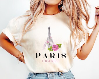 May include: Cream-colored t-shirt featuring a graphic of the Eiffel Tower with pink accents and a pink rose. The text "PARIS FRANCE" is printed below the graphic in black and pink.