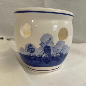 May include: A white ceramic candle holder with a rounded shape and a blue painted landscape scene featuring a windmill. The vessel has two circular cutouts on the sides and a blue rim around the top edge, showcasing a classic delft-style design.