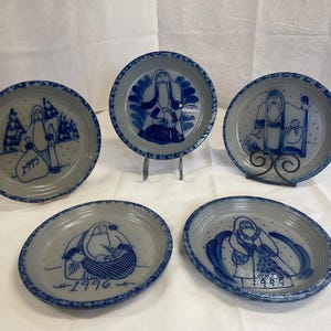 Puede incluir: Un conjunto de cinco platos redondos de cerámica con base gris claro y diseños pintados a mano en azul oscuro. Cada plato presenta una ilustración única de una figura, y algunos muestran los años 1995, 1996 y 1998 en tinta azul.