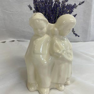 May include: A cream-coloured ceramic vase shaped like two figures standing side by side, wearing hats and traditional clothing. The hollow top of the vase is filled with a bunch of dried purple lavender stems, adding a vibrant contrast to the smooth, glossy finish of the off-white ceramic material.