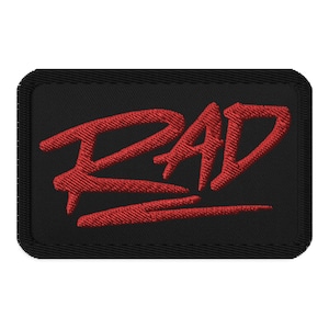 May include: Black rectangular patch with the word "RAD" embroidered in red. The letters are stylized and bold, with a slight slant. The patch has a black border.
