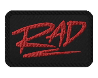 May include: Black rectangular patch with the word "RAD" embroidered in red. The letters are stylized and bold, with a slight slant. The patch has a black border.