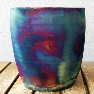 Puede incluir: Una maceta de cerámica hecha a mano con un esmalte iridiscente. La maceta presenta una mezcla de tonos azules, morados y verdes, creando una apariencia única y artística. La forma cilíndrica se asienta sobre una superficie de madera.