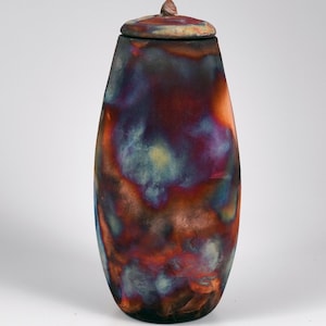 Può includere: Un'urna alta in ceramica con coperchio. L'urna ha una forma allungata e ovale, ricoperta da una glassa con una miscela di colori, tra cui rossi profondi, viola, blu e tocchi di giallo. Il coperchio ha un piccolo puntale appuntito.